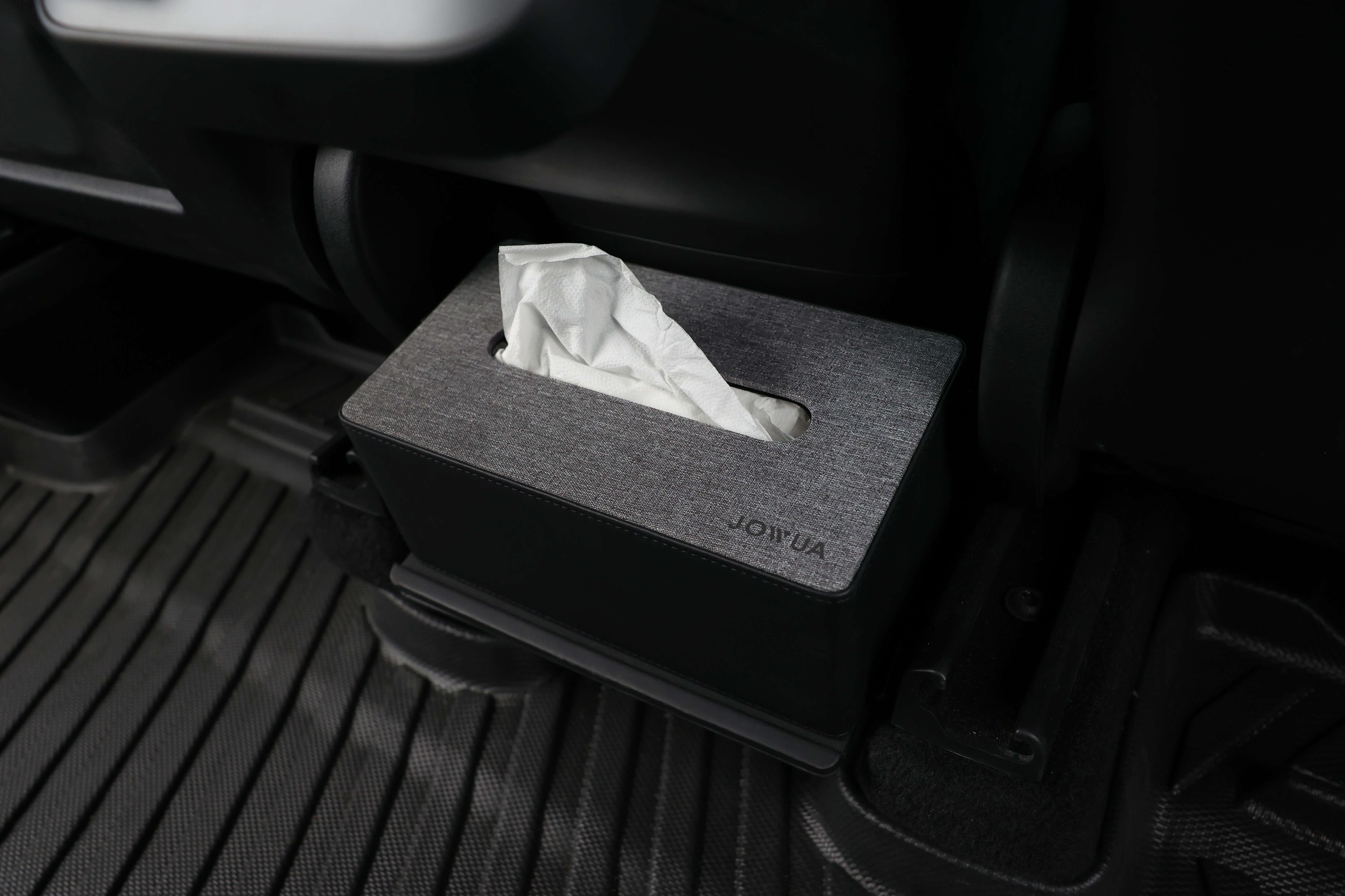 A JOWUA Tissue & Storage Box, crafted from premium materials with a sleek black fabric cover, is positioned on the floor of a car between the driver's seat and the center console. A single tissue is partially pulled out, complementing the stylish interior and floor mat.