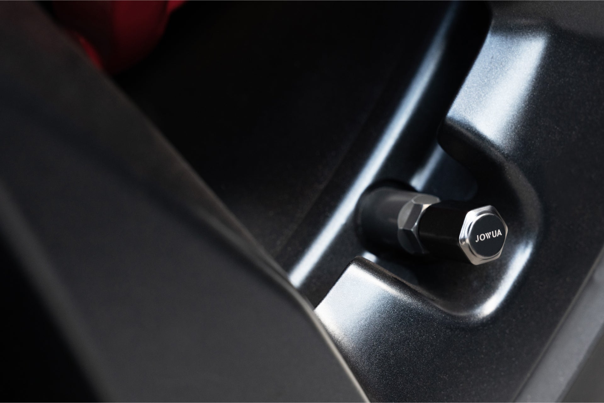 Close-up of a black JOWUA tire valve cap, made from zinc alloy, nestled in a vehicle's tire wheel well, providing airtight leak protection amidst smooth, dark plastic panels.