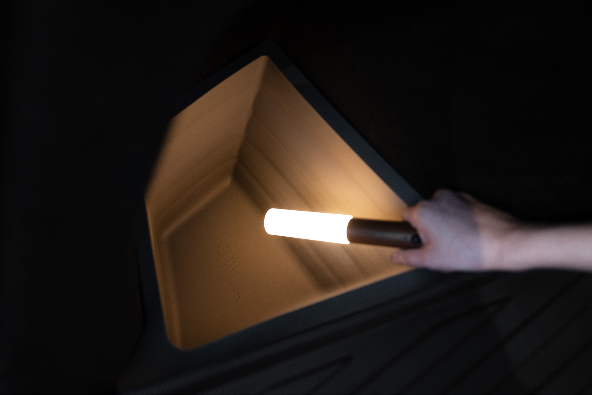 A hand holding a lit, cylindrical light brightens the interior of a dark, empty trunk compartment with textured walls, ideal for uncovering hidden gems in your Model Y using the JOWUA Trunk Storage Bins Bundle.