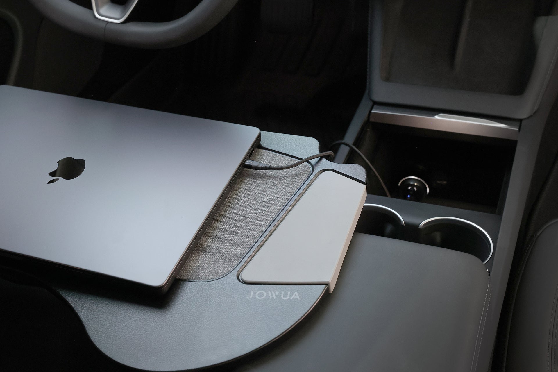 A MacBook and smartphone are connected with cables to a JOWUA 165W PD3.1 Car Charger, placed on the car's front seat center console, near cup holders, using a desk tray optimized for Tesla vehicles.