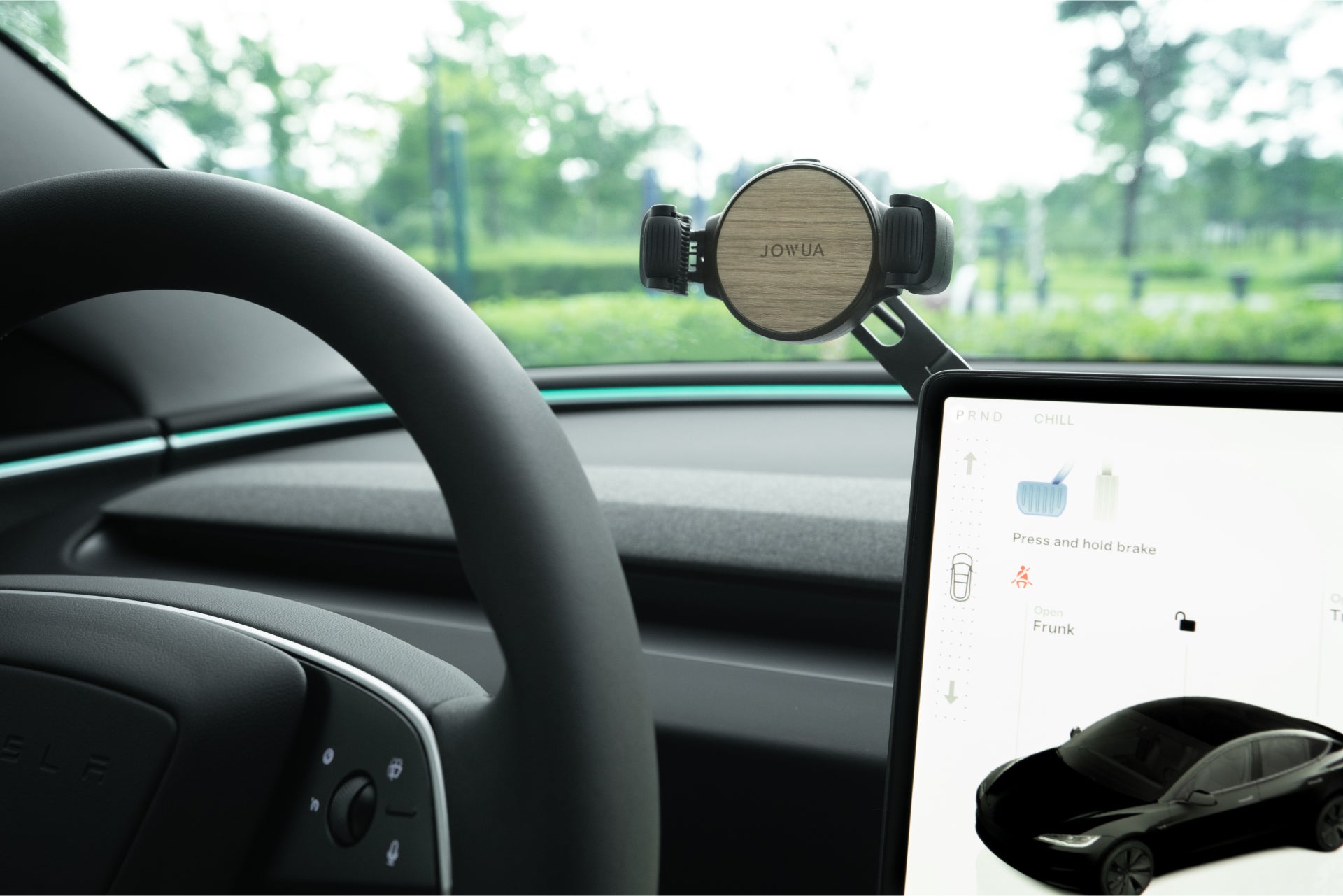 A close-up of a Tesla dashboard shows the JOWUA 6D Tesla Compatible Invisible Foldaway Car Mount Qi2 Wireless Charger near the steering wheel and a large touchscreen on the right, with greenery visible through the windshield.
