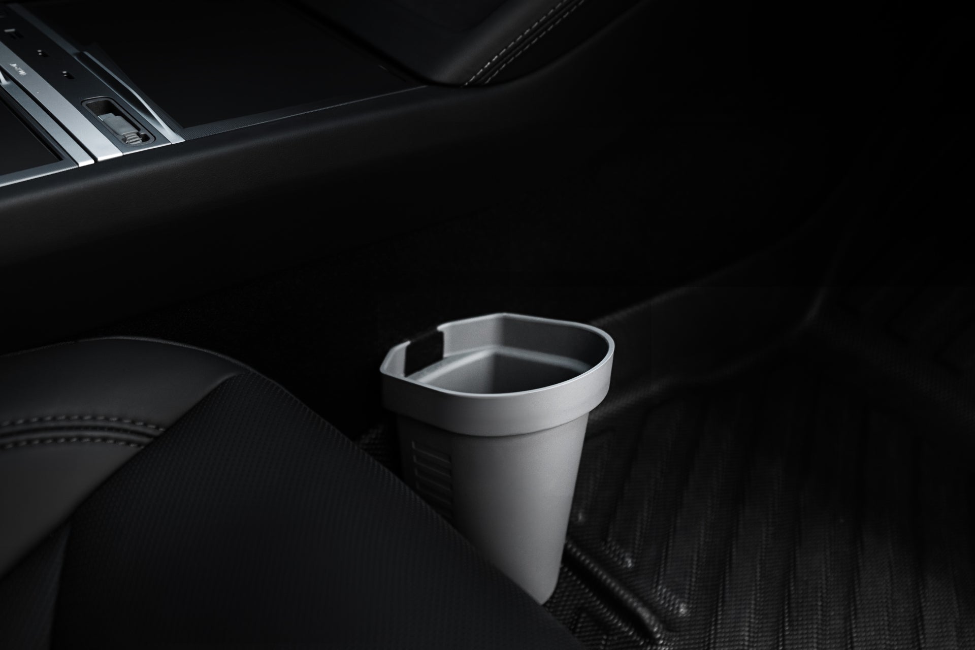 Close-up of a Tesla Model's interior featuring a gray, octagonal storage box made of sleek silicone material resting on a black floor mat. It's situated near the passenger seat, with part of the dashboard visible above, under dim lighting that accentuates its modern elegance. This is one of the Door Side Storage Boxes from JOWUA.