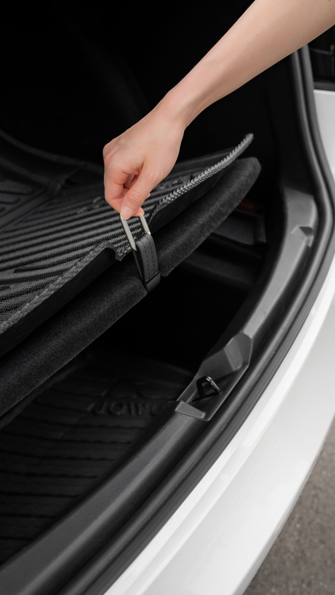 A hand lifts the floor mat in the trunk of a white Tesla Model 3, revealing an easily accessible sub-trunk compartment beneath the sleek black interior, compatible with the JOWUA Snap Hook for Tesla Model S/3.