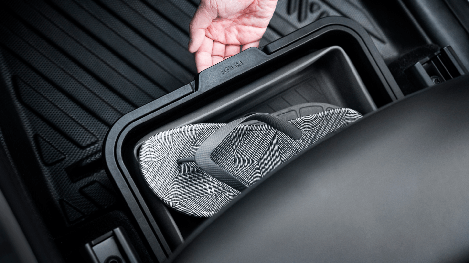 A hand lifts the lid of the Under Seat Collapsible Organizer for Tesla Model X Y by JOWUA, revealing a pair of black and white patterned flip-flops inside. The car's interior surrounds the organizer, showcasing its foldable and expandable design for multipurpose use.