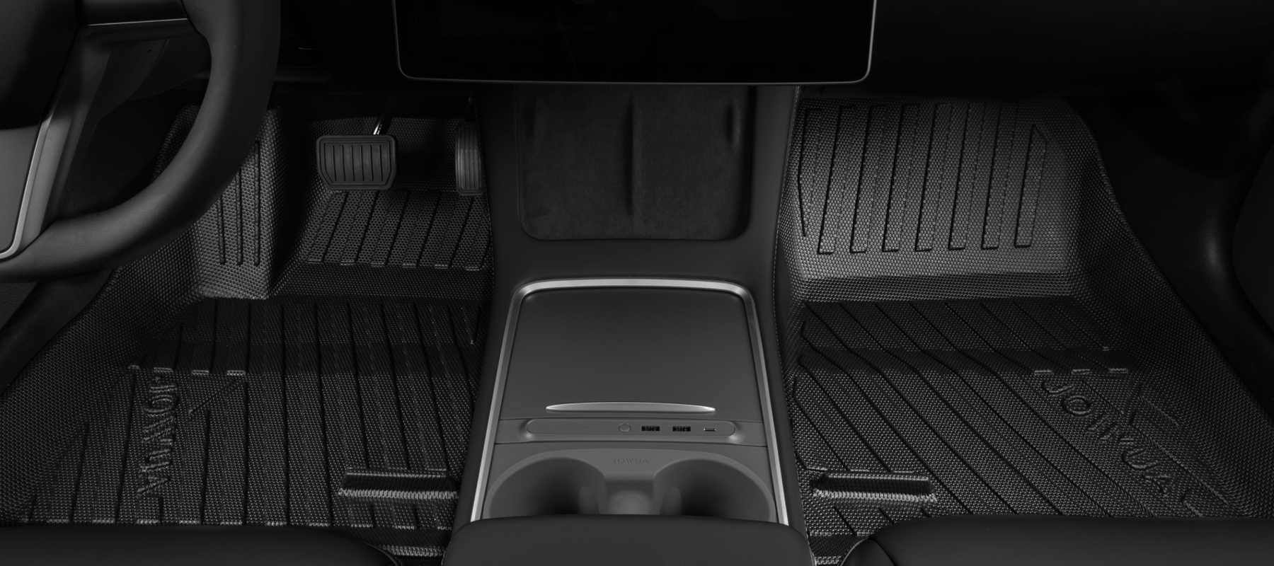 Close-up view of a car's interior, showing the driver's seat area. Visible are the steering wheel, pedals, central console, and textured floor mats, all in a sleek black design.