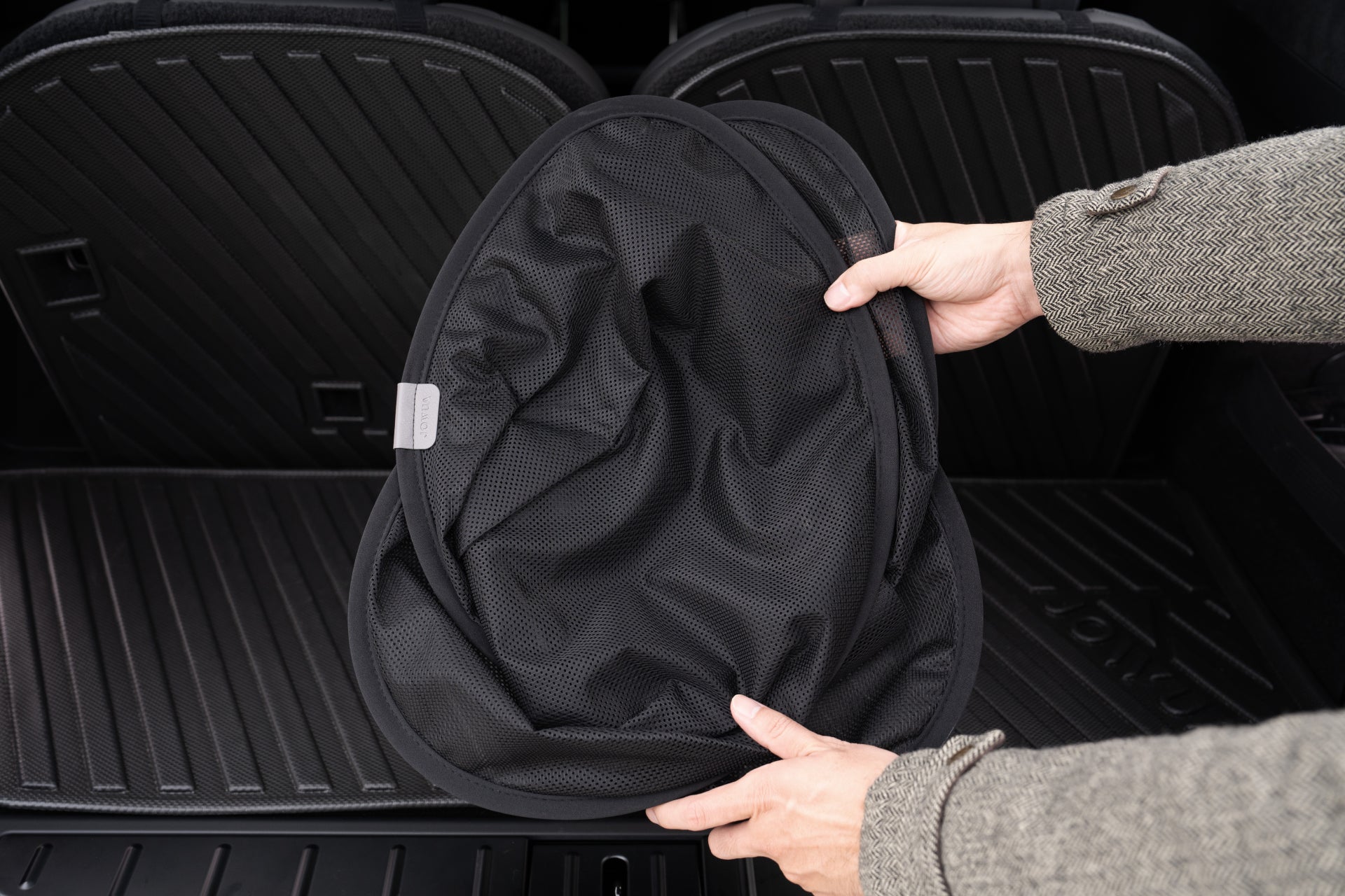 A person holds the JOWUA Rear Lift Gate + Triangular Window Sunshade for Model X above a car's trunk with rubber floor mats. The sunshade, crafted from high-density woven fabric, is folded to highlight its compact and portable design.