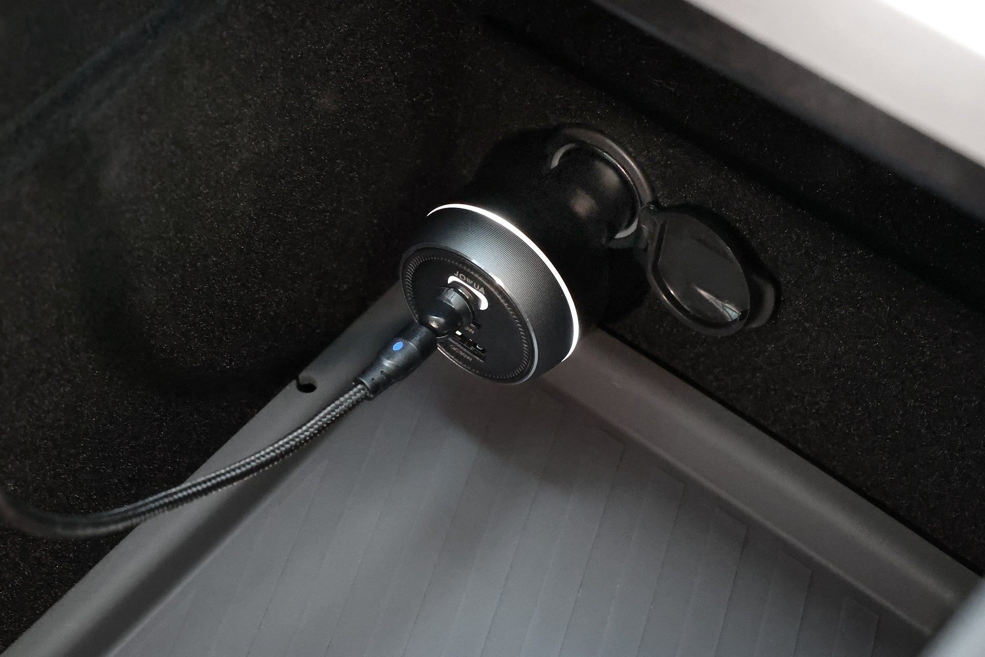 A close-up of the JOWUA Portable Air Compressor + Charging Solution Kit plugged into a car’s 12V outlet, delivering high-speed charging via USB, with part of the car’s interior floor and side panel in view.