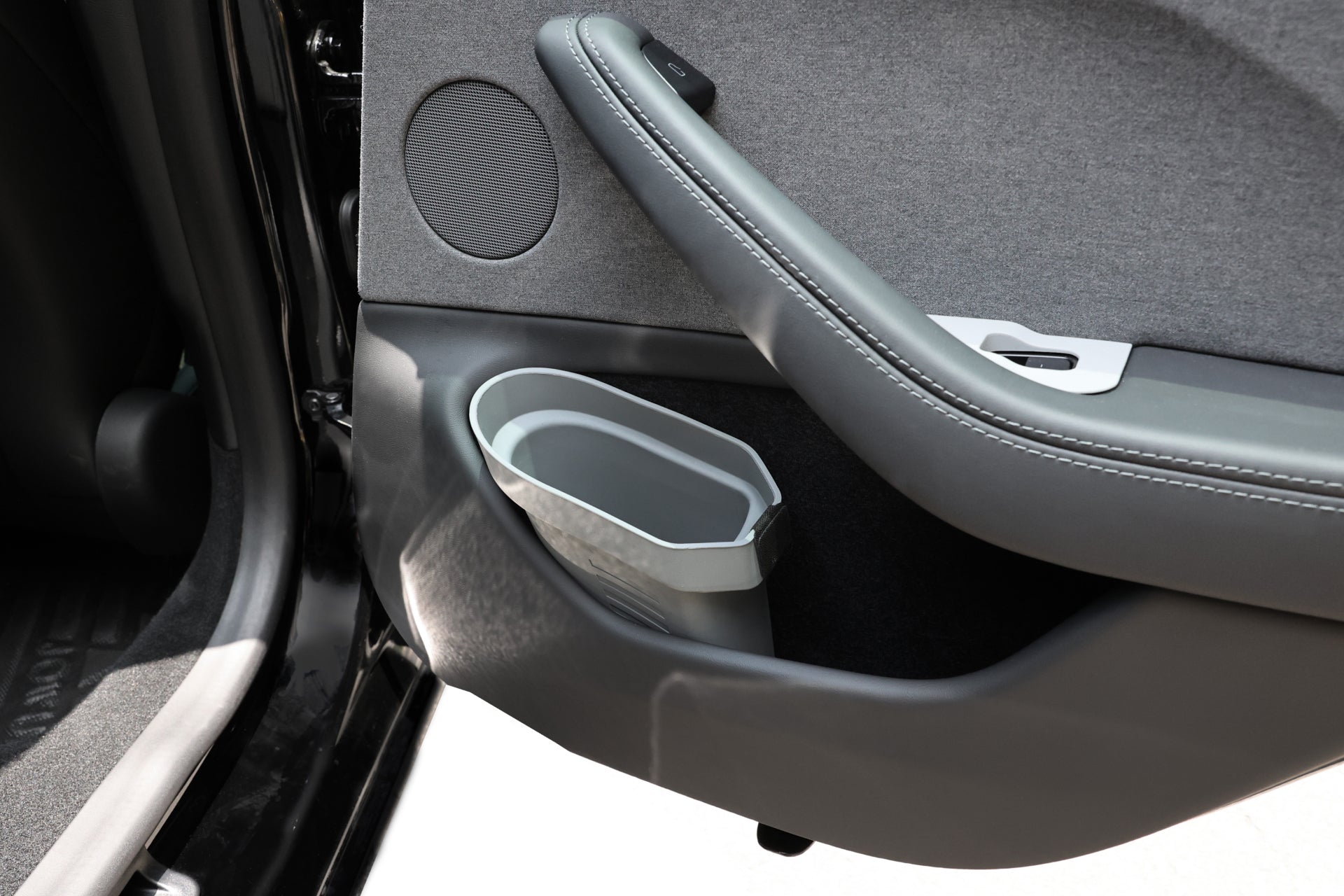 Close-up of the JOWUA Door Side Storage Box in a Tesla Model’s car interior features a grey fabric panel, a speaker, and an integrated cup holder with a geometric design. The padded handle showcases stitched detailing, complemented by the sleek storage bin crafted from durable silicone material.