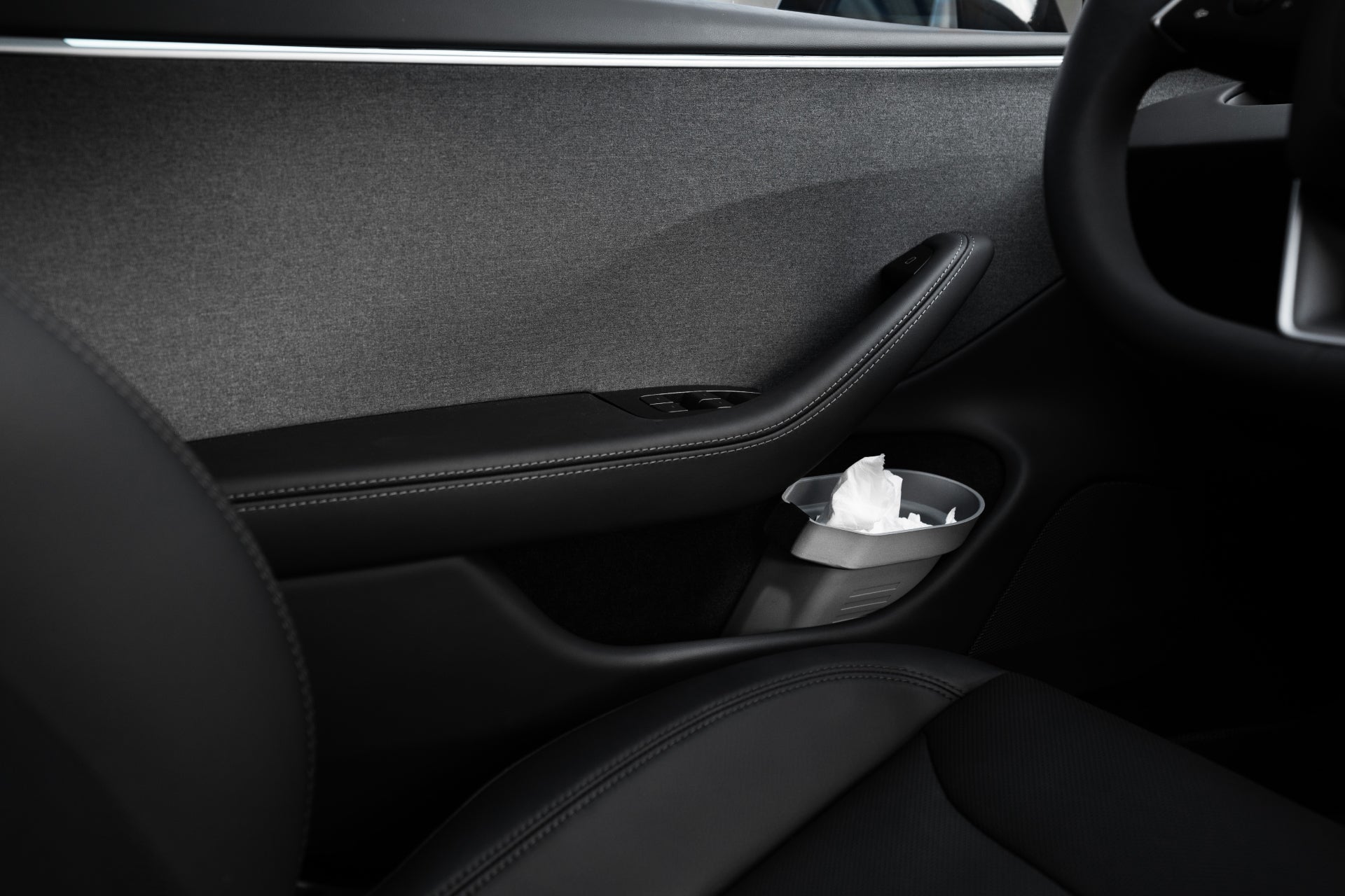 The interior of a Tesla Model car door highlights a sleek, dark design with a black armrest, window controls, and JOWUA's silicone material cup holder that includes a tissue. The gray upholstery enhances the modern and minimalistic aesthetic while smartly integrating JOWUA’s Door Side Storage Box (Set of 2) for added convenience.