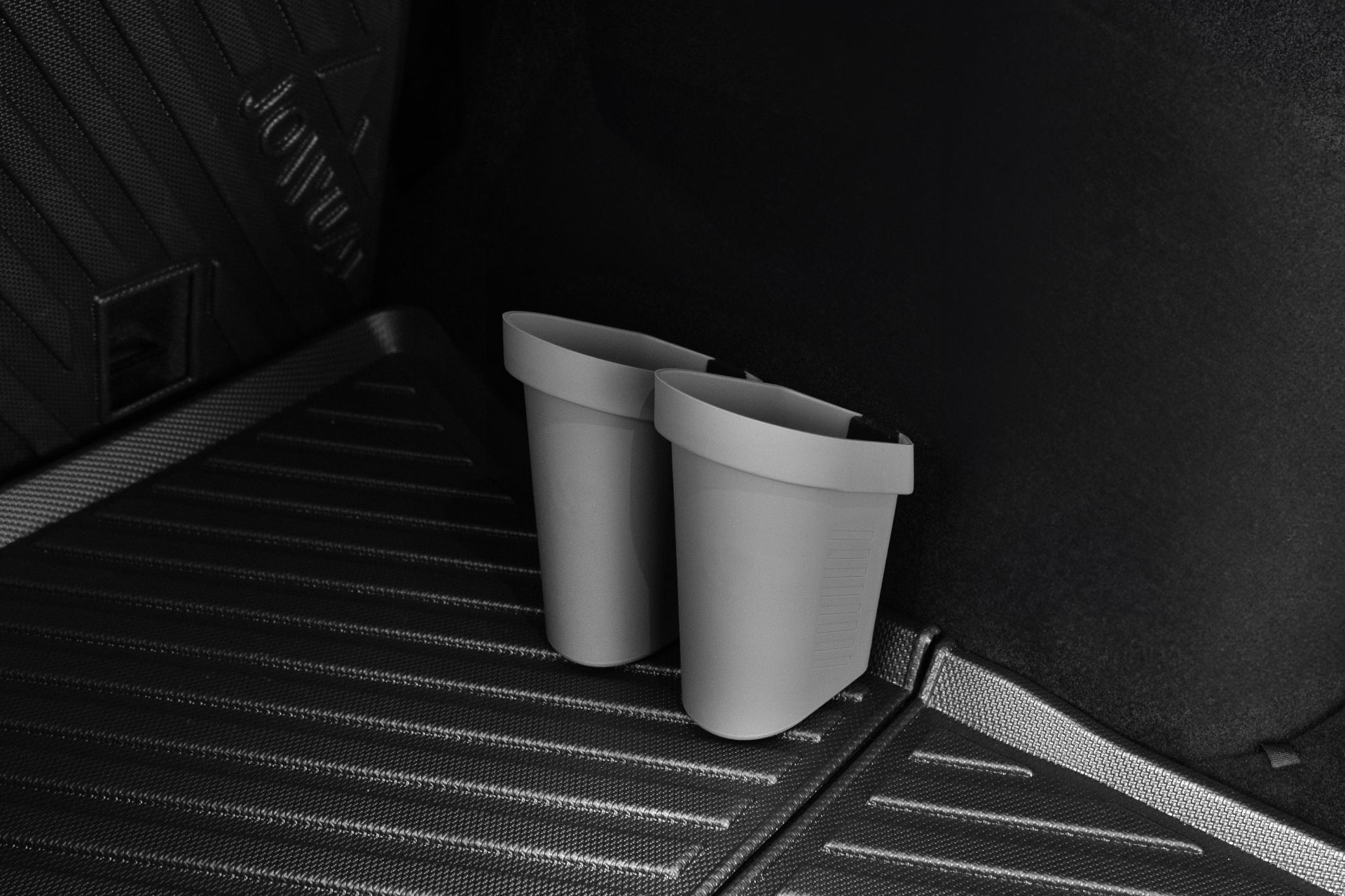 Two JOWUA Door Side Storage Boxes, which are gray and cylindrical in shape and made from silicone material, are placed next to each other on a textured, dark car floor mat. This mat, designed for Tesla Models, features parallel grooves and a raised border with light elegantly reflecting off its surface.