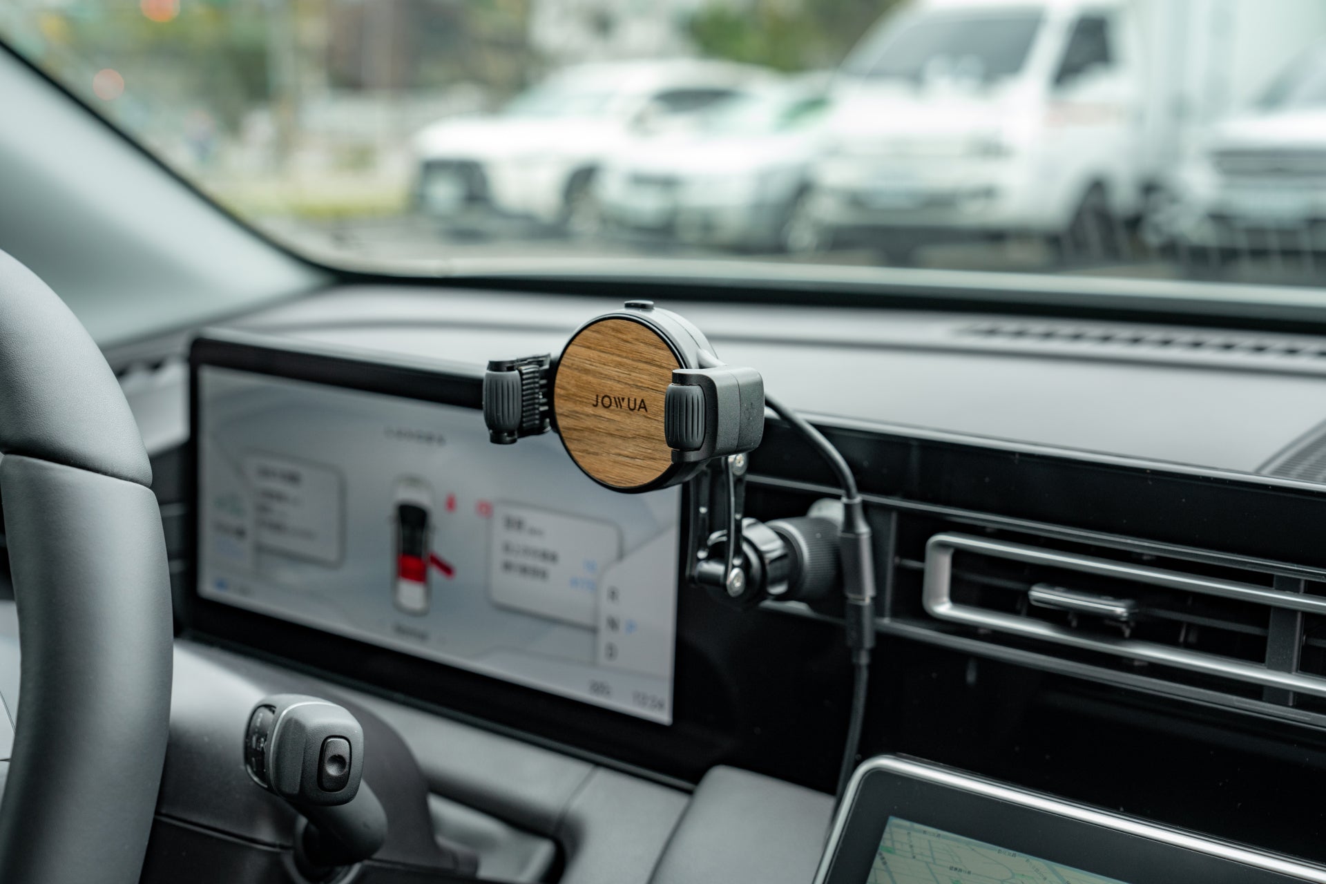 A car interior showcases a JOWUA Universal Air Vent Car Mount (Qi2 Universal Roller Mount) attached to the dashboard. A touchscreen with navigation details is visible behind, and through the windshield, several cars are seen nearby, emphasizing wireless charging capabilities.