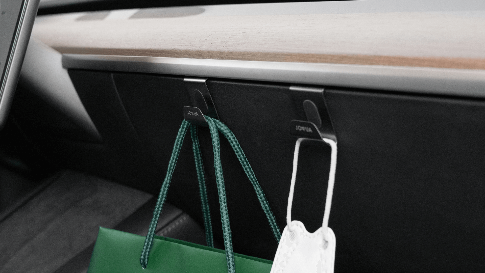 Two JOWUA Glovebox Hooks, seamlessly attached to a Tesla Model 3's sleek dashboard, elegantly hold a green shopping bag and a white face mask. Complementing the organized interior, a subtle wooden trim design meets the stainless steel accents.