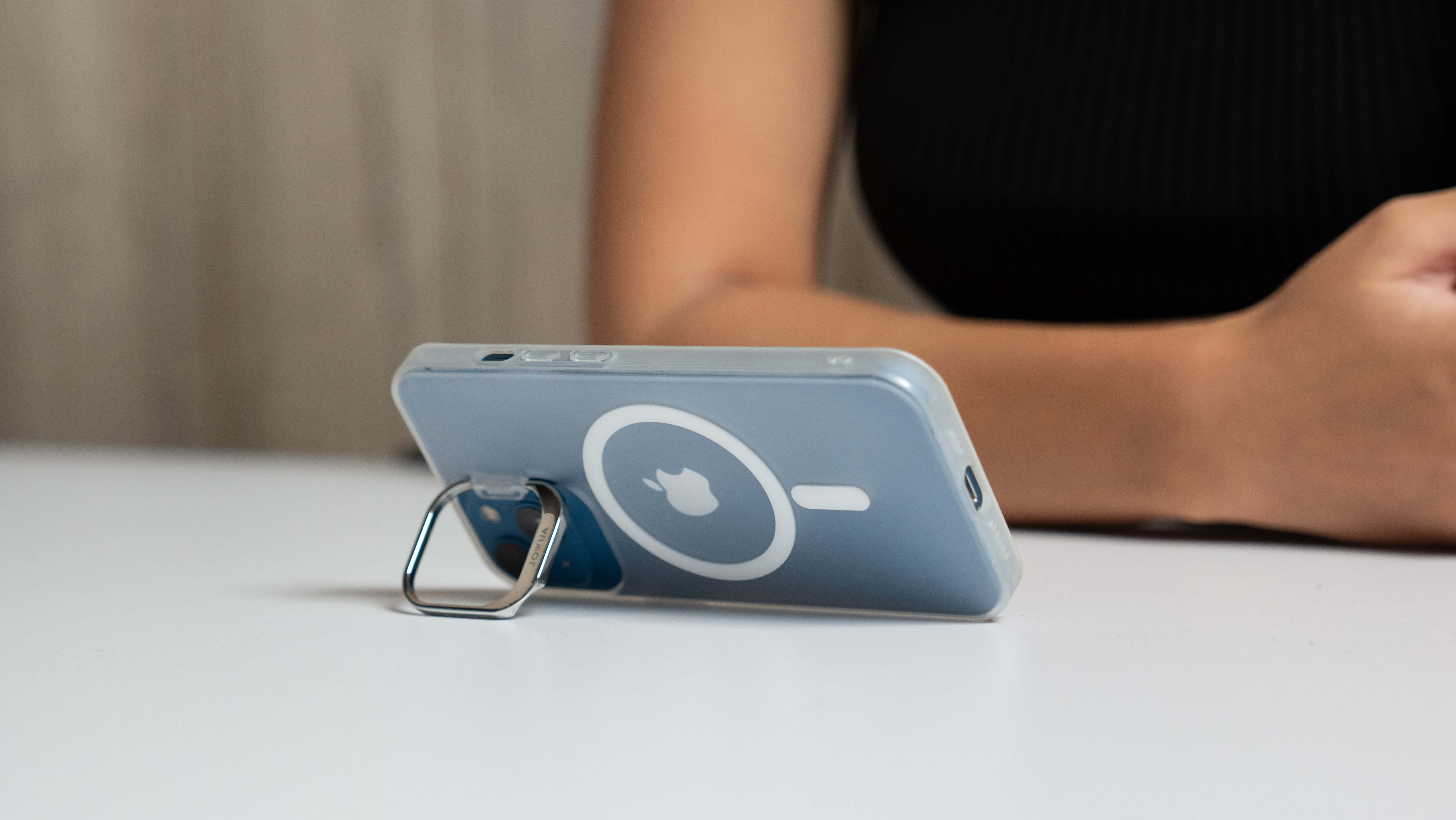 A person sits at a white table, holding a smartphone with a light blue JOWUA iPhone Case for Tesla Car Mount featuring MagSafe compatibility and a kickstand design. The phone, positioned horizontally on the stand, complements their black sleeveless top with its sleek style.