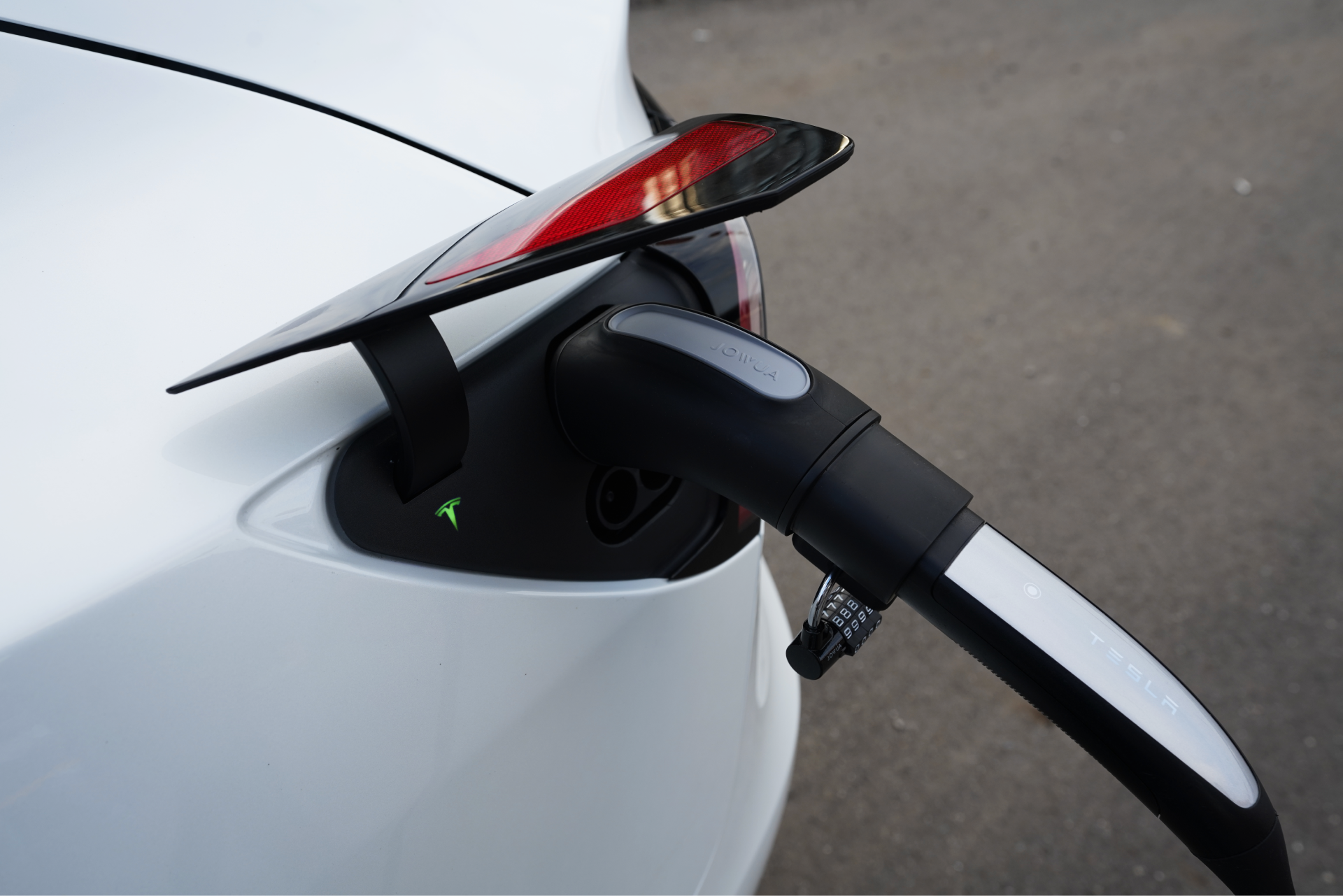 A close-up view captures a white electric car being charged, featuring the JOWUA TPC(NACS) to CCS2-AC (TYPE2) Adapter connected to the vehicle's port. The tail light and part of the rear can be seen against a blurred asphalt backdrop, accentuating the sleek padlock-inspired design.